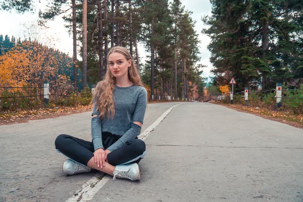 Uma linda garota loira sentada no meio de uma estrada vazia i — Fotografia de Stock