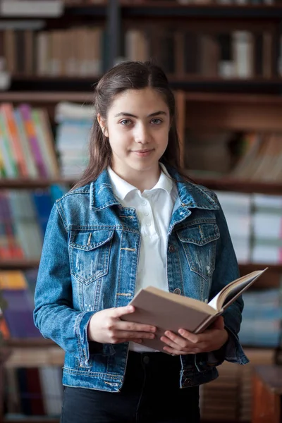 Bela estudante de pé na biblioteca e lendo um livro — Fotografia de Stock