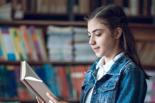 Bela estudante sentada na biblioteca e lendo um livro — Fotografia de Stock