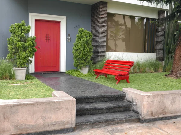 Horizontal view of a gray and red color mint condition small house with a red wooden door and a red wooden bench located in Miraflores district of Lima. In the image there is the garden