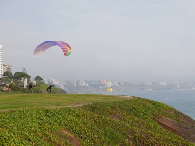 Lima, Peru. October 23, 2017. A pilot in a no engine multi colored paraglider is ready to jump and fly from Raymondi park located over the cliff on the coast of Miraflores touristic district of Lima clipart