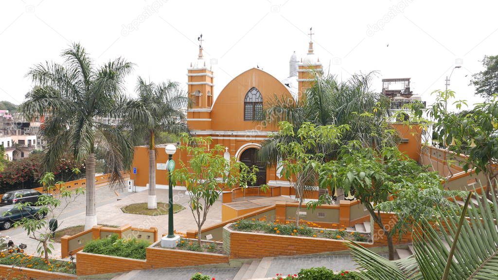 Scenic view of a yellow and white color hermitage facade and garden at different levels in the main square point of Barranco beatnik district of Lima. In the image there are also trees, a street, cars, a staircase, plants, flowers and lampposts 