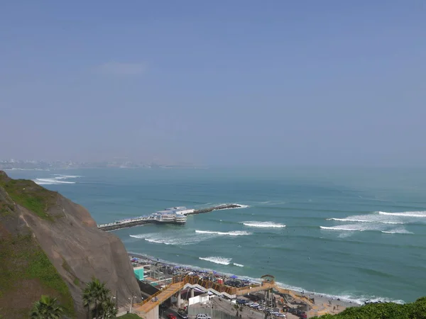 Scenic view to the Pacific Ocean and a pier with a built construction from the top of a Lima cliff in El Parque del Amor located in Miraflores district of Lima. There is a blue sky day with horizon over calm ocean.