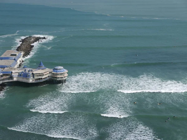 Scenic view to the La Rosa Nautica restaurant built over a pier at coastline of Miraflores district of Lima. The photo was taken from El Parque del Amor over a cliff on a blue sky day. Unrecognizable surfers are in the image.