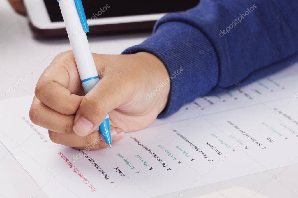 student's hand holding blue pen taking English test on white desk in class
