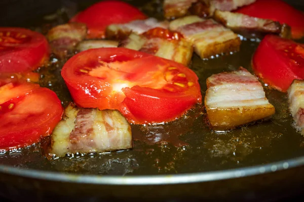 Red sliced tomatoes and sliced bacon are fried in metal frying pan. Close-up.