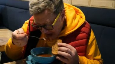 Man in bright clothes eats soup from meat, vegetables, beet and tomatoes - borscht with sour cream. With a metal spoon in the cafe or restaurant. Close-up.