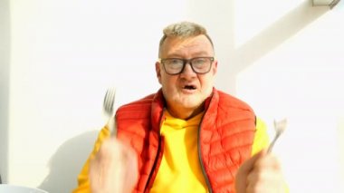 Yaşlı adam parlak giysiler içinde beyaz milliyet Oynaşman, çatal ve kaşık ile oynarken, sipariş edilen yiyecek için bekleyen, bir restoran ya da Kafe oturan.