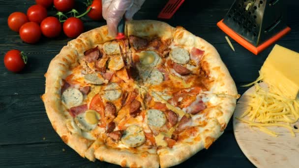 Las manos cortan la pizza, por los trozos, con la salchicha, el tomate y el queso, con el cuchillo especial - la rueda del cortador. En la mesa de madera también hay tomates y queso rallado . — Vídeos de Stock