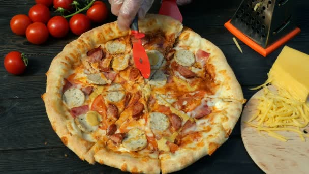 Las manos cortan torpemente la pizza, por los trozos, con la salchicha, el tomate y el queso, con el cuchillo especial - la rueda del cortador. En la mesa de madera también hay tomates y queso rallado . — Vídeos de Stock