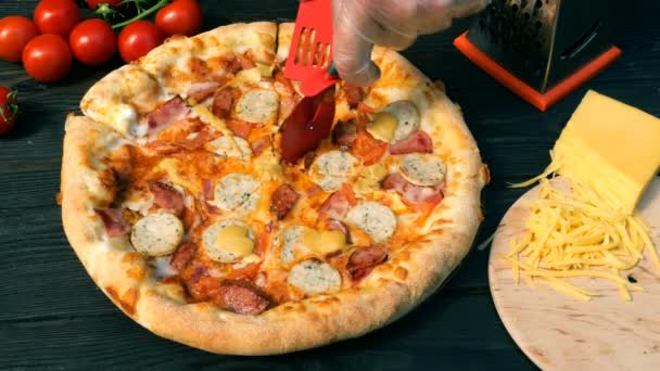 Руки незграбно відрізають піцу, на скибочках, з ковбасою, помідорами та сиром, зі спеціальним ножем - фрезою. На дерев'яному столі також є помідори і тертий сир . — стокове відео