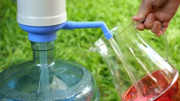 Die menschliche Hand füllt einen durchsichtigen Krug mit kaltem Wasser aus einer großen Flasche oder einem Kühler mit einer Wasserpumpe. vor dem Hintergrund des grünen Rasens. Selektiver Fokus. im Freien. — Stockvideo