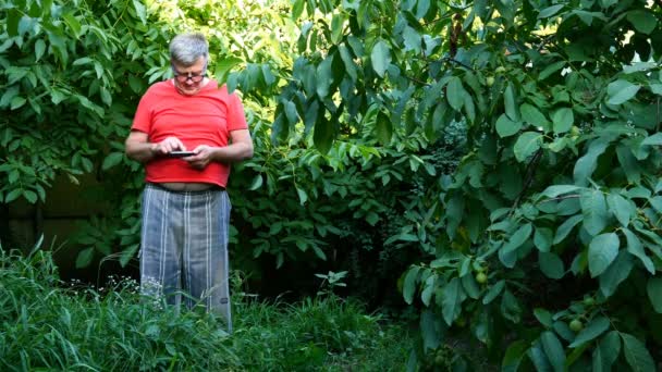Hombre mayor en ropa casual, tal vez un pensionista, utiliza su teléfono inteligente en el patio trasero o en el jardín . — Vídeos de Stock