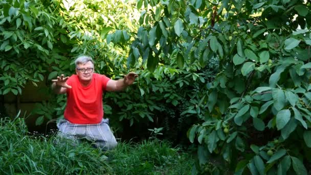 Senior man i casual kläder, kanske en pensionär, gör knäböj på bakgården eller i trädgården. Använd som koncept hälsosam livsstil för medelålders människor. — Stockvideo