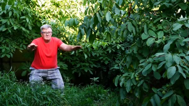 Senior man in casual clothes, perhaps a pensioner, doing squats in the backyard or in the garden. Use as a concept healthy lifestyle for middle-aged people. — Stock Video