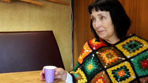 Senior woman, caucasian ethnicity, dressed in poncho drinks coffee from disposable cup while sitting in cafe and talk something. Average plan. — Stock Video