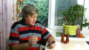 Kafkas kökenli yetişkin bir erkek, günlük giysiler içinde, el kol hareketi yapıyor ve duygusal olarak, habercisiyle, akıllı telefonuyla, odanın veya mutfağın penceresinin yanındaki masada oturuyor. Ortam planı.