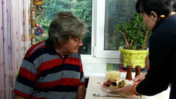 Mujer adulta madura, etnia caucásica en ropa casual están sirviendo comida para su marido en la mesa de comedor cerca de la ventana en la habitación. Masculino comiendo comida con placer. Una cena familiar. Plan medio . — Vídeos de Stock