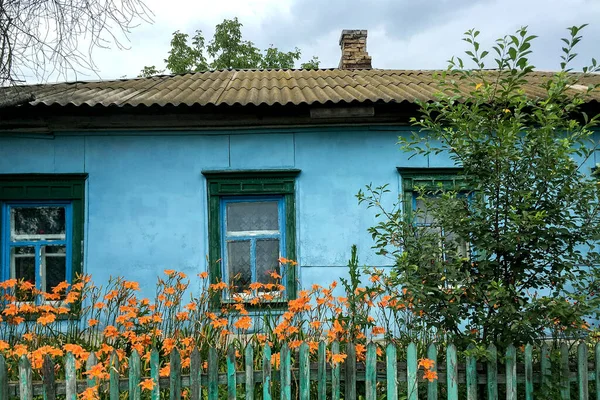 Old blue tidy house with wooden windows and tiled roof in village or countryside. Behind wooden fence. The concept of a calm everyday life. Selective focus. Close-up. Outdoors.