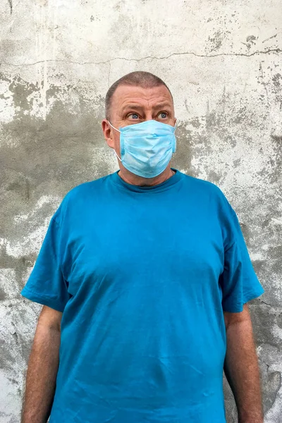 Short-haired caucasian ethnicity mature man in blue t-shirt with medical mask on his face to protect COVID-19, looking to side against background of gray plastered wall. Vertical shot. Close-up.