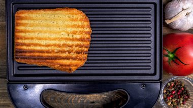 Çizgili altın hamurlu ızgara beyaz ekmek, açık siyah sandviç makinesinde, ahşap kırsal masada yatıyor. Baharat, domates, sarımsak. Hızlı yemek pişirme. Üst Manzara. Düz yatıyordu. Yakın plan. Kapalı.