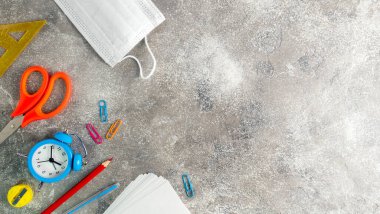 Farklı kırtasiye malzemeleri, mavi alarm saati ve tıbbi maske. Coronavirus zamanında okul malzemeleri. Evde öğreniyorum. Okul konseptine geri dönelim. Üstü açık. Düz yatıyordu. Uzayı metin için kopyala. Yakın plan..