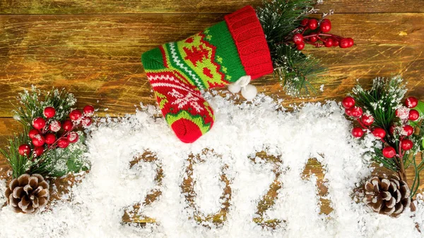 Christmas branches with berries and cones in decorative snow, which says 2021 and knitted Christmas stocking or Santas boot. Christmas mood concept. Top view. Flat lay. Close-up. Festive.