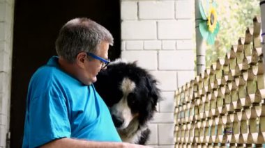 Yaşlı yetişkin bir adam evin verandasında otururken Bernese Dağ Köpeğini okşayıp kucaklıyor. Evcil hayvanlar için sevgi ve arkadaşlık kavramı. Yakın plan. Açık havada..