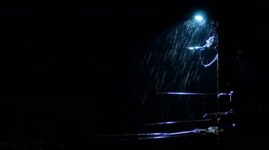 Gece ya da karanlık akşam. Şiddetli yağmur püskürtüleri yanan sokak lambasının arka planında. Yıldırımlar köyde ya da kasabada evleri aydınlatıyor. Orta boy..