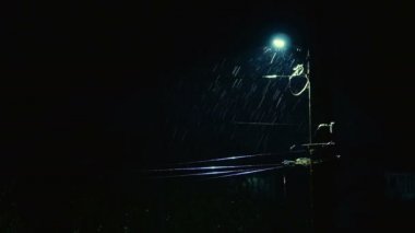 Gece ya da karanlık akşam. Şiddetli yağmur püskürtüleri yanan sokak lambasının arka planında. Yıldırımlar köyde ya da kasabada evleri aydınlatıyor. Orta boy..