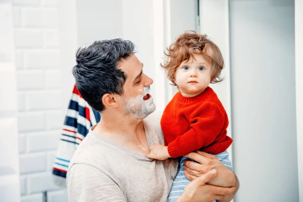 中年父亲早上抱着幼儿阳光 早上在浴室刮胡子 — 图库照片