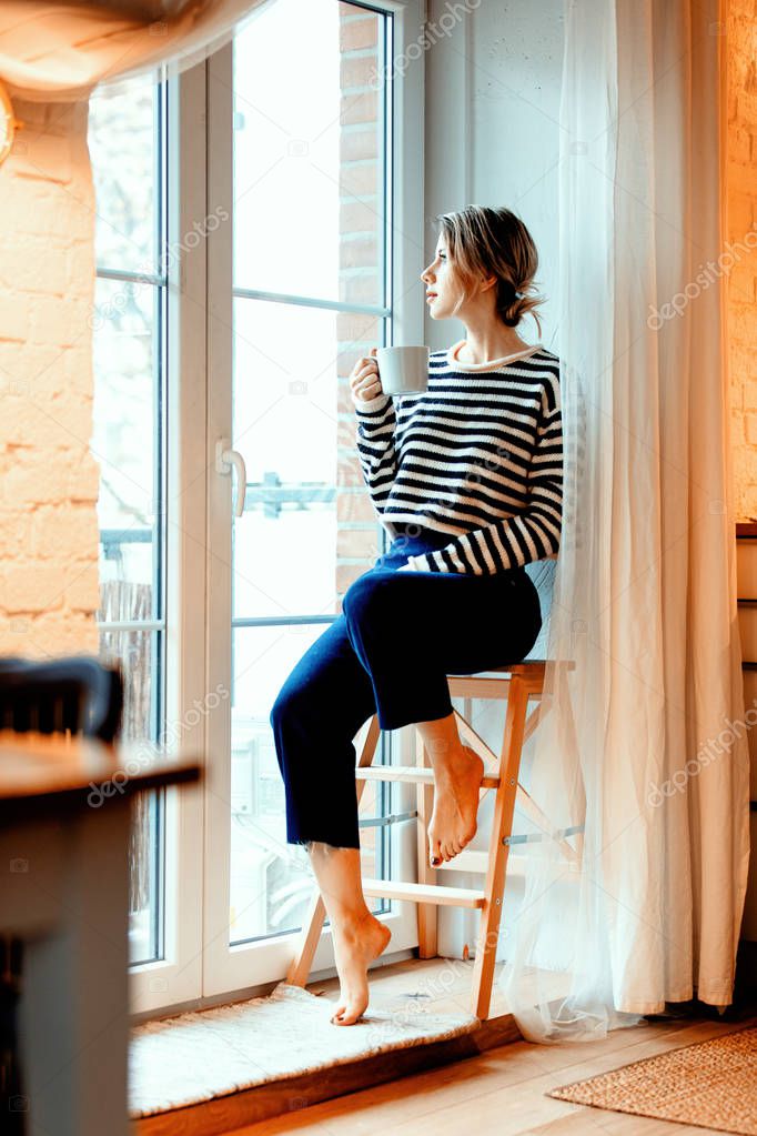 beautiful redhead girl with cup of coffee is sitting on little stairs at home near window.