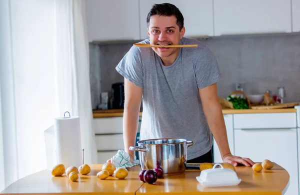 Young man standing with spoon in mouth cooking at kitchen.