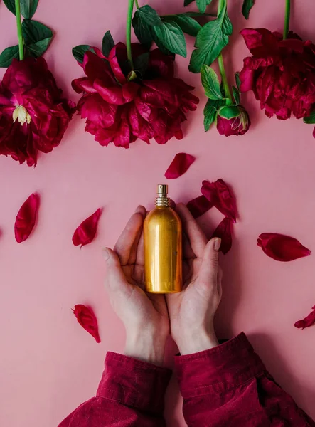 Female hands holding golden perfume bottle near peonies flowers