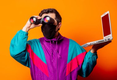 Yüz maskeli stil adamı ve 80 'lerin eşofmanı, dizüstü bilgisayarı ve turuncu arka planda dürbünü var.