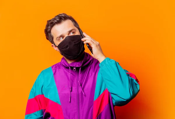 Style man in face mask and 80s tracksuit with mobile phone on orange background