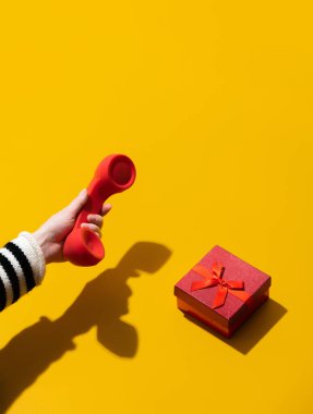 Kadın el, sarı arka planda hediye kutusunun yanında kırmızı cep telefonu tutuyor. 