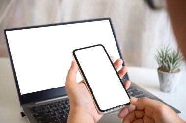 Ev ofisinin çalışma masasında boş beyaz ekran akıllı telefon ve boş beyaz laptop ekranı olan bir el. Sanat çalışmanız için akıllı telefon ve bilgisayar ekranı oluşturun.