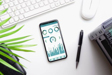 Ofis masasının en üst görüntüsü, iş raporları grafiği beyaz masadaki akıllı telefondan. Mobil uygulama kavramları yoluyla işletme yönetimi.