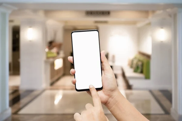 Mockup blank white screen smartphone hand pointing and touch mobile phone isolated on white with blurred background of beautiful living room, blank screen cellphone mock up for your  advertisement.