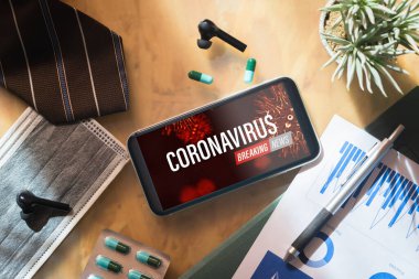 Coronavirus ya da Covid-19 Salgını Son Dakika Haberleri arka plan güncelleme kavramı. İş adamının ahşap masasında yüz maskeleri ve tıbbi tabletleri olan model cep telefonu..