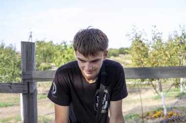 Genç çocuk, kahverengi saçlı, gülümseyen, fotoğrafçı, siyah tişört, tahta çitin yanında duruyor, kamera kayışı, boynunda, arka planda genç bir bahçe, çiftlik, açık hava, gündüz, kırsal alan, yaz, yeşillik, yüz, açık hava, çocuk