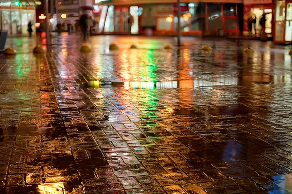 Rainy night in the city. Wet street, colored lights reflection and blurred silhouettes with umbrellas.