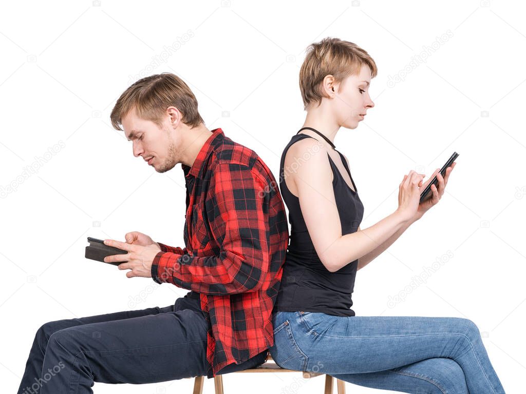 Young couple sitting on one chair back to back and looking at his smartphones. Isolated on white. Gadget addiction, millennials, hipsters, loneliness together and modern communication problems theme