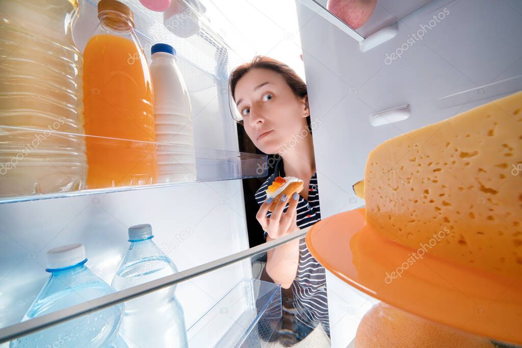 Tricky girl holding sweet cake and looking into fridge full of different food. Wide angle view from inside 