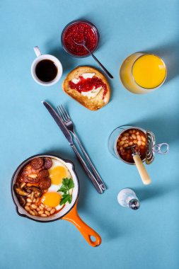 İngiliz kahvaltısı bir pişirme tavada kızarmış yumurta, sosis, fasulye, kızarmış ekmek ve kahve mavi masada düz lay