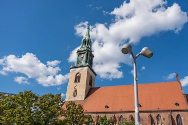 Berlin 'deki St. Mary Kilisesi mavi gökyüzünün altındaki kulesiyle yükseliyor. Bulutlar ve ağaçlar sahneyi oluşturur.