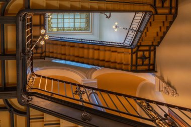 Aşağıdan görülen süslü merdiven, vitraylı tavan penceresine çıkıyor. Lambalar zarif yapıyı aydınlatır.