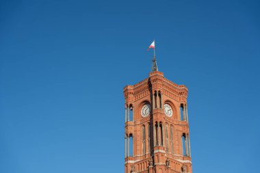 Kırmızı tuğlalı Rotes Rathaus kulesi açık mavi gökyüzünün altında uzun duruyor. Bayrak en üstte gururla dalgalanır.