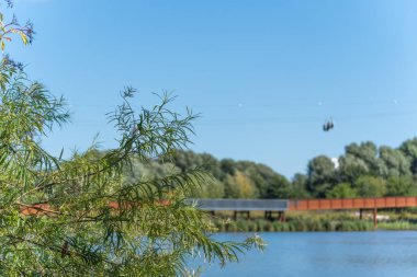 Yeşil dallar modern bir köprüyle bir göl manzarası oluşturur. Bir teleferik açık mavi gökyüzünü geçiyor.